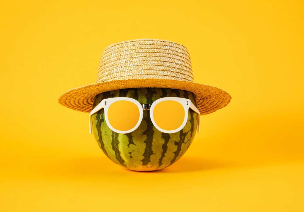 Eine Wassermelone vor einem gelben Hintergrund trägt einen Sonnenhut und eine Sonnenbrille mit gelben Gläsern. Das Bild symbolisiert Spaß und Vielfalt und steht im Zusammenhang mit den fröhlichen Events von International Friends e.V. .